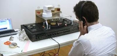 Another view of Jon Beaton\'s sound and smell machine. The main element is a machine that looks like a record player with scientific measuring beakers on top. The user is seen sitting in front of the machine wearing headphones.