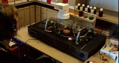 Jon Beaton\'s installation in use. The main element is a machine that looks like a record player with scientific measuring beakers on top. The user is seen sitting in front of the machine wearing headphones.