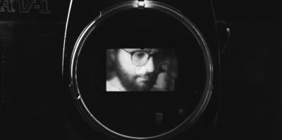 An analogue photo camera with the lens removed, revealing the camear\'s mirror and filmmaker Jon Beaton\'s face reflected in that mirror.