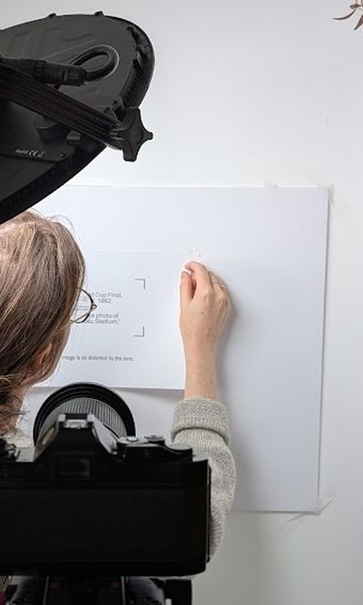 A woman affixing a presentation slide to a white wall in order for it to be photographed by the camera in the foreground.