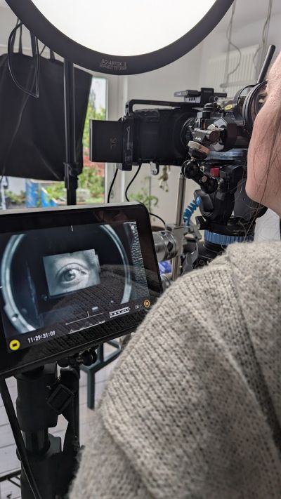 A woman looking into the viewfinder of an analogue photo camera that is being filmed by a digital cinema camera.