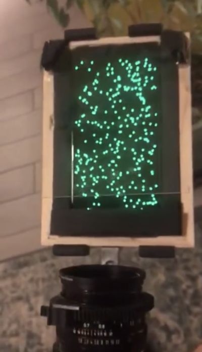 A top-down view of a wooden shelf attached from below the camera lens. The shelf holds a mobile phone facing up. On the screen are lots of glowing green dots. Above the screen is a piece of transparent acrylic at a 45 degree angle.