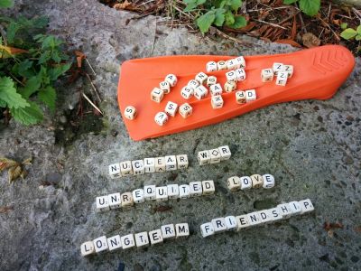 A bright-orange flipper is laying on the ground upside-down. Scattered on the flipper are a load of dice with letters on them instead of numbers. Some of those letter cubes have been arranged on the ground beside the flipper to form the words: Nuclear War, Unequired Love and Longterm Friendship