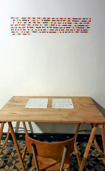 A white wall covered in colourful squares of tape arranged in a neat pattern. Below is a makeshift desk with two pieces of paper on it.